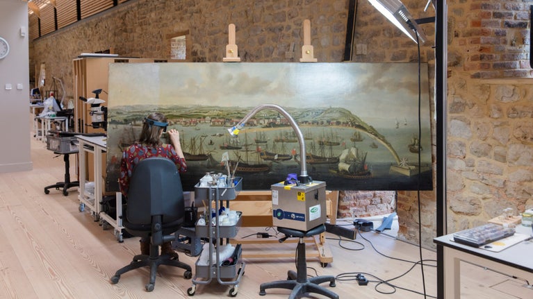 A conservator works on A view of the port of Bridgetown, Barbados with extensive shipping oil painting from Dyrham (Gloucestershire) in The Royal Oak Foundation Conservation Studio, Knole, Kent